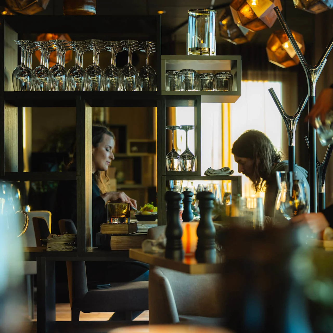 Bord med middagsgäster njuter av måltider i en varmt upplyst restaurang, omgiven av vinglas och hyllor, medan en servitör handskas med drycker.
