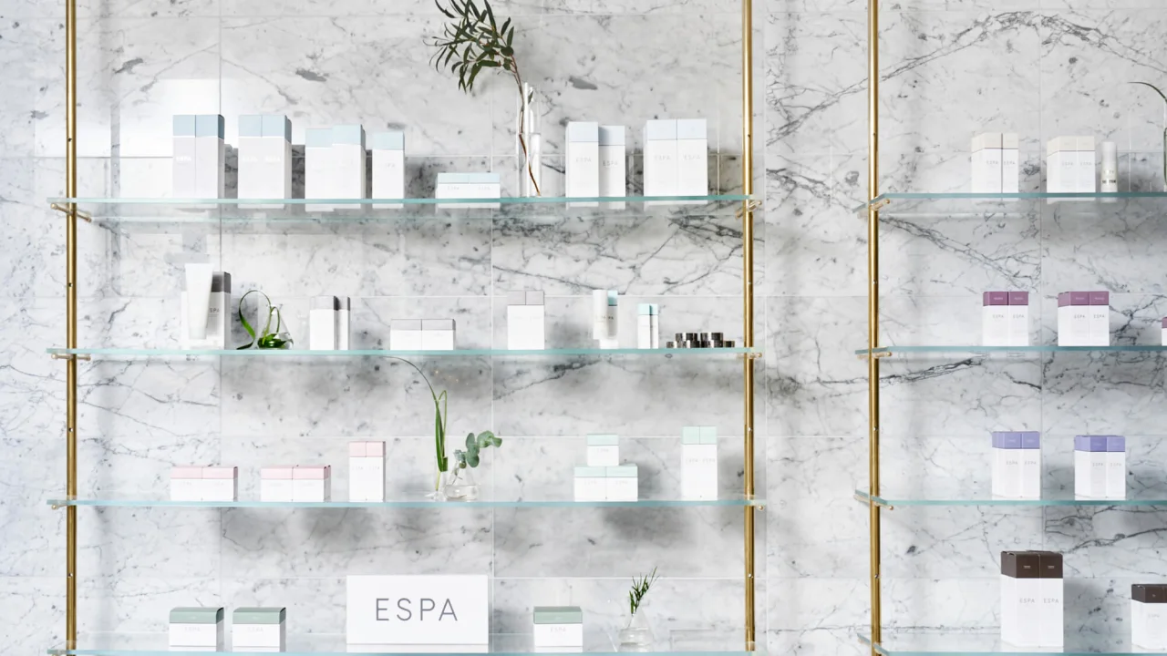 Glass shelves with cosmetic products against a marble wall. A sign reads 'ESPA'.