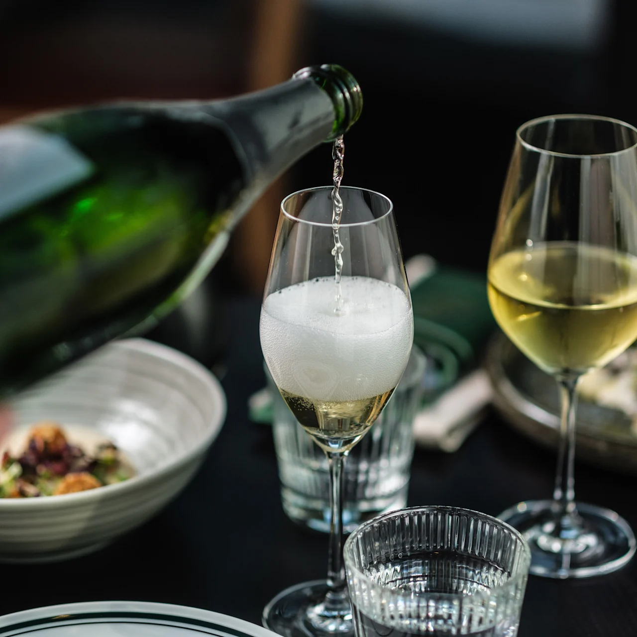 En grön flaska häller mousserande vin i ett glas, skapar skum. Bredvid finns ett fullt vinglas. Tallrikar och en servett syns på ett mörkt bord.