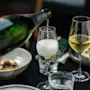 En grön flaska häller mousserande vin i ett glas, skapar skum. Bredvid finns ett fullt vinglas. Tallrikar och en servett syns på ett mörkt bord.