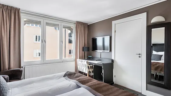 A bedroom with a made bed, window curtains open, television, desk, and mirror, suggesting a hotel room.