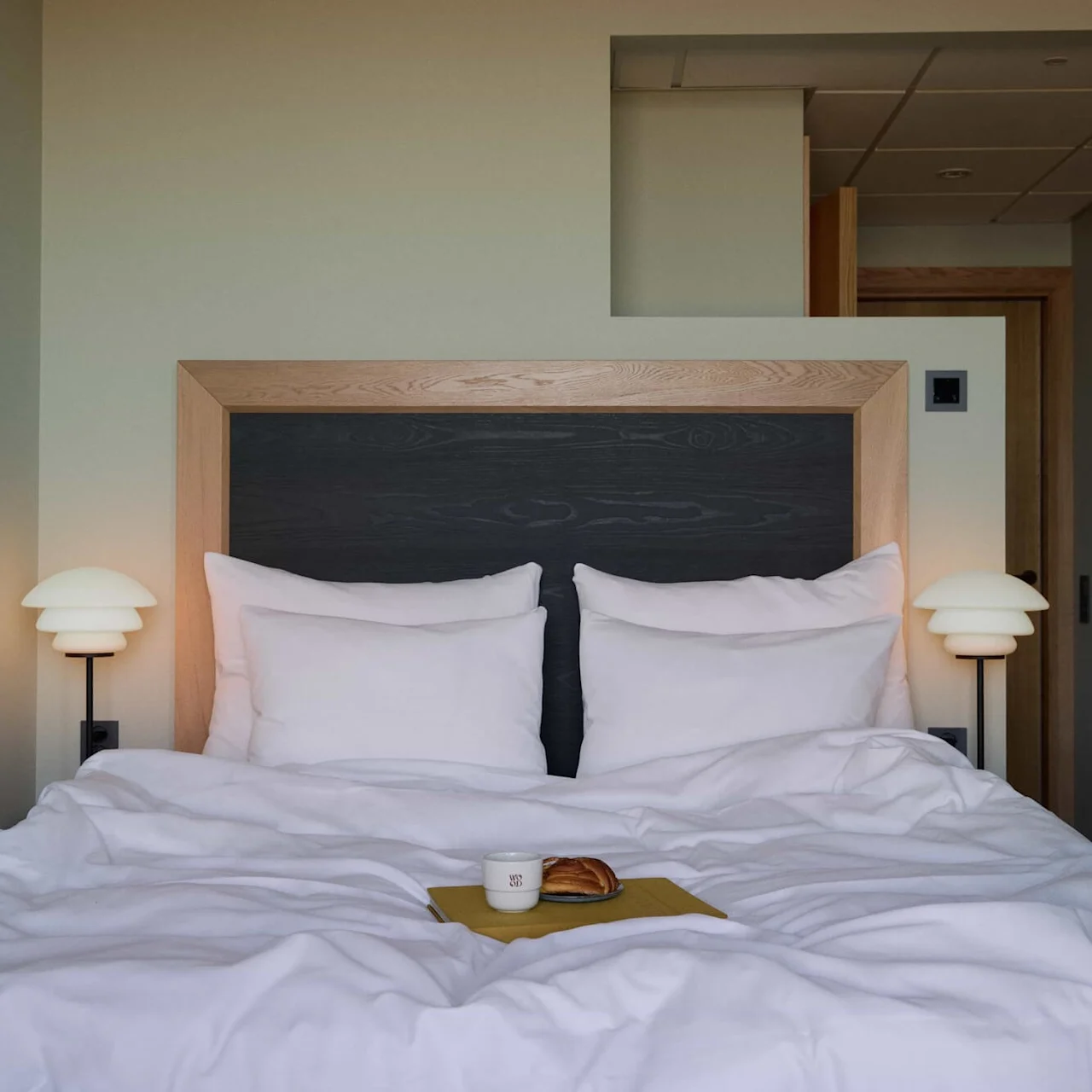 A neatly arranged bed with white pillows and lamps sits in a minimalistic room. A coffee cup and croissant are on a napkin. Text on the cup reads "MB."
