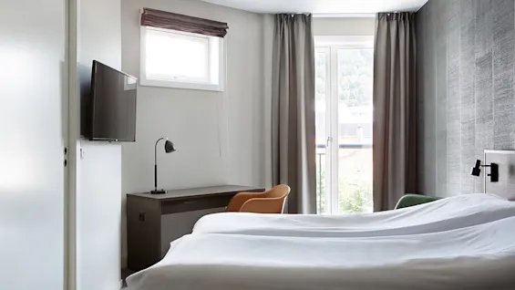 Seng pænt redt med hvidt sengetøj, placeret ved siden af et træbord og lampe i et lyst rum. En vægmonteret tv og vindue med gardiner giver en hjemlig atmosfære.
