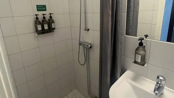 Shower area with three soap dispensers mounted on tiled wall, a showerhead above, and a gray curtain. Beside, a sink with a soap dispenser; mirrored tiles reflect the setting. Text reads: "ORORI MENUSY パタザ"