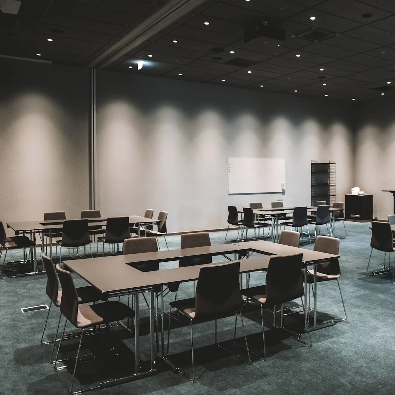 Bord och stolar arrangerade i små grupper i ett rymligt, mattbelagt konferensrum med dämpad belysning. En whiteboard och ett podium står mot väggen.