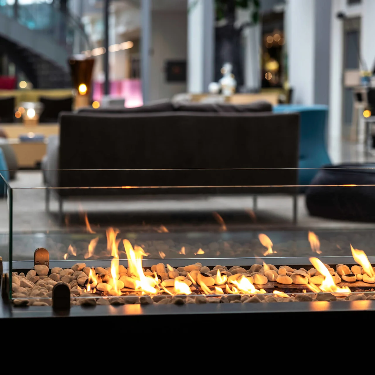 En glass-innbygd peis med flammer og småstein brenner innendørs. Den står i et moderne salongområde med stilige møbler og en trapp i bakgrunnen.