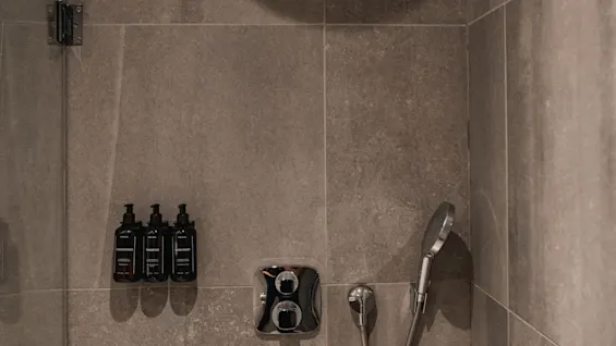 A modern shower with grey tiles, a rain shower head, and wall-mounted bottles for CONDITIONER, SHAMPOO, BODY WASH.