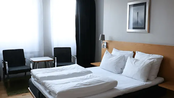 En seng med hvitt sengetøy er pent oppredd i et hotellrom, flankert av to stoler og et lite bord under to lyse vinduer.