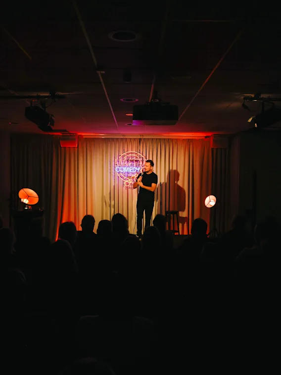 A stand up comedian performing on a dimly lit stage in front of a seated audience.