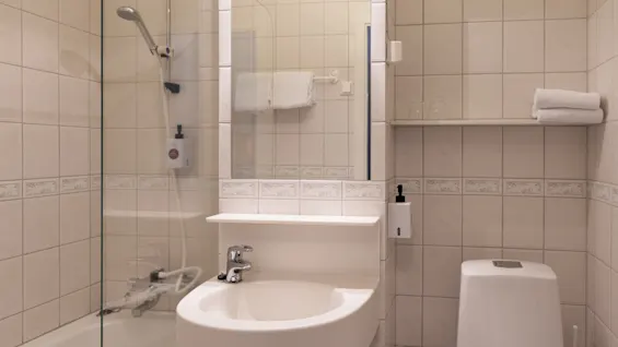 En vit vask med en kran är placerad bredvid en glasinkapslad dusch i ett kaklat badrum. En toalett finns intill och en hylla har vikta handdukar och toalettartiklar.