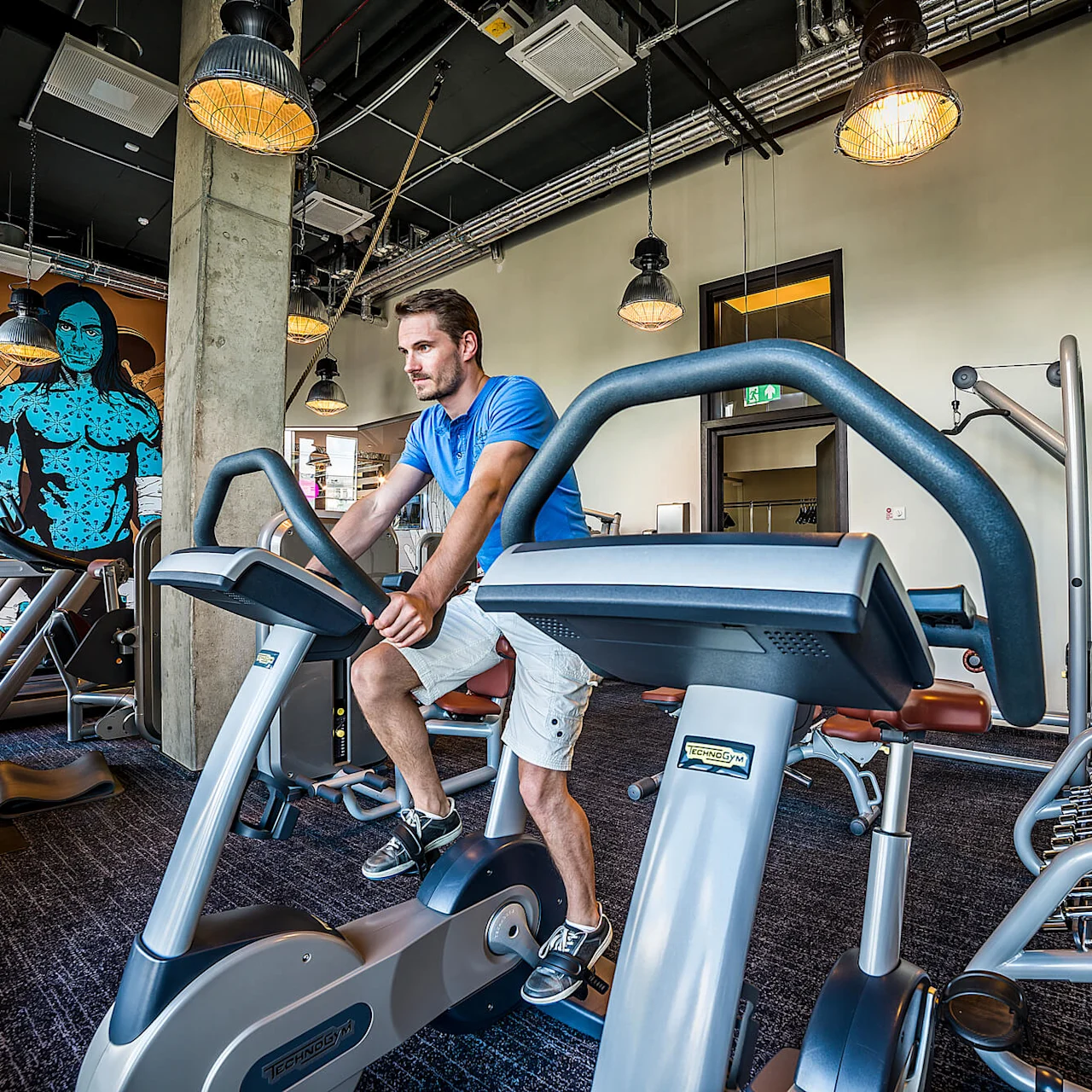 Man cyklar på motionscykel i modernt gym, omgivet av vikter och utrustning. Färgglad väggkonst lyder, "MY ROCK & ROLL IS BETTER THAN YOUR BODYBUILDING."