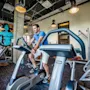 Man cycling on stationary bike in modern gym, surrounded by weights and equipment. Colorful wall art reads, "MY ROCK & ROLL IS BETTER THAN YOUR BODYBUILDING."