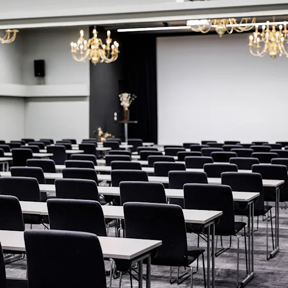 En stor konferansesal satt opp i klasseromsoppsett med rader av sorte stoler, lange bord, lysekroner og et lerret i front.