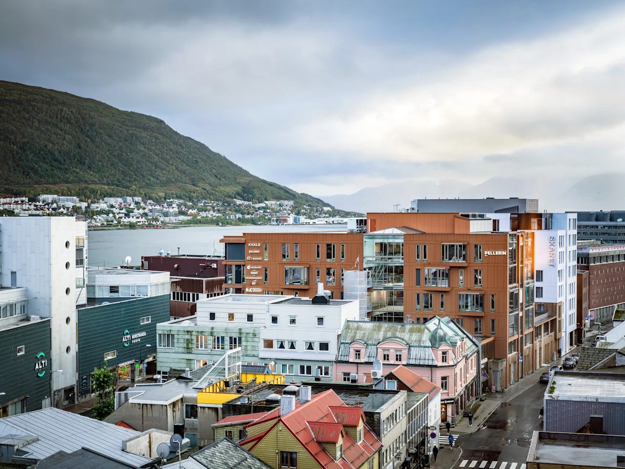 Kaupunkikuva rakennuksineen veden äärellä, vuoria taustalla pilvisen taivaan alla. 