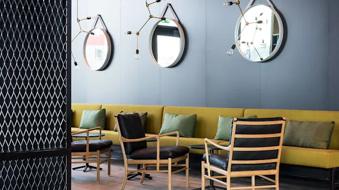 Elegant chairs in a lounge setting face a modern couch with green pillows, framed by blue walls adorned with mirrors and unique light fixtures. A patterned divider partially shows the space.