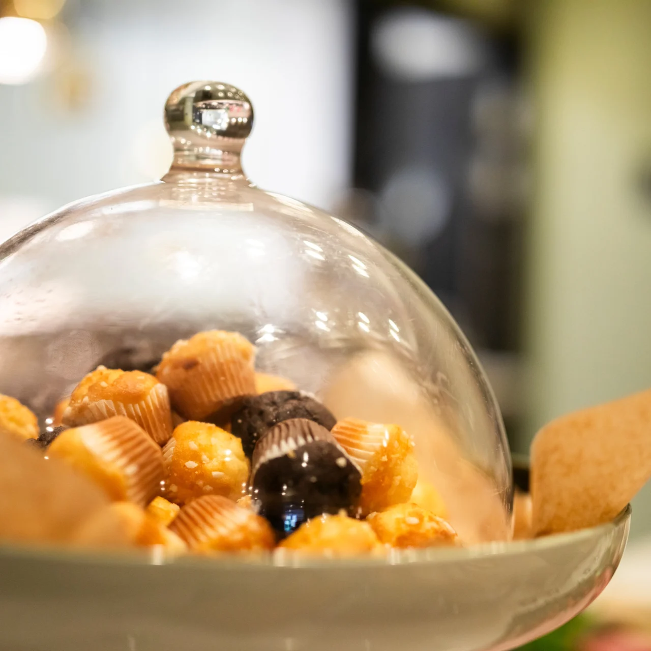 Glass dome covers assorted chocolates on a plate, resting on parchment paper in a cozy kitchen setting with blurred background.