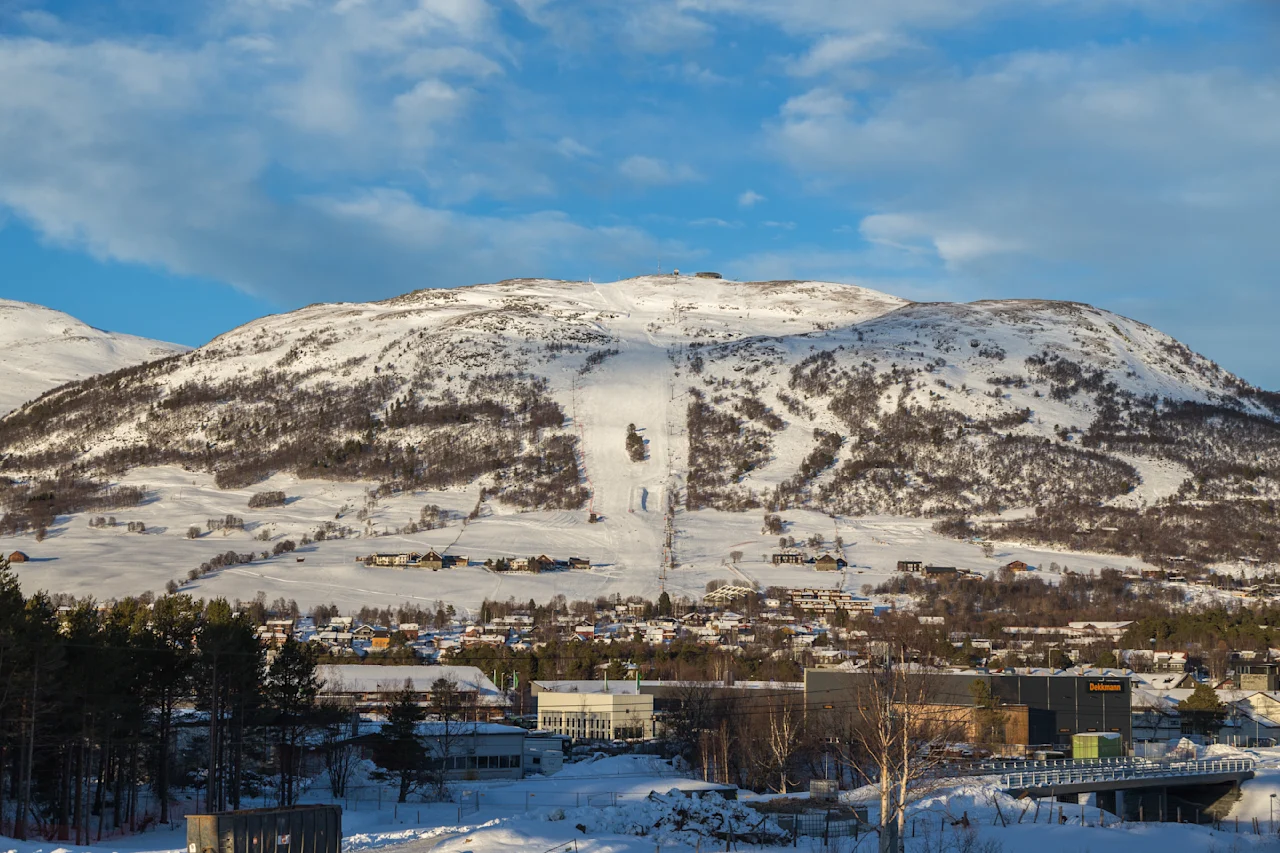 Snødekte fjellskråninger med skiløyper strekker seg ned mot en landsby med spredte hus og bygninger, omgitt av skoger under en delvis skyet blå himmel.