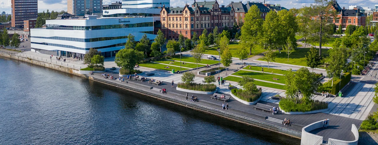 Aerial view of Umeå's vibrant waterfront, with people enjoying the promenade and green spaces, framed by diverse city architecture.