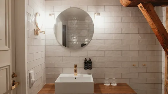 Et stilrent badeværelse med hvid flisevæg, et rundt spejl, en moderne vask og varme trædetaljer.