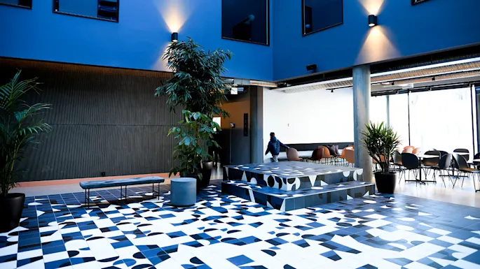 A geometric-patterned floor stretches through a modern, blue-walled interior space with potted plants and seating areas. A person walks past tables and chairs under bright overhead lighting.