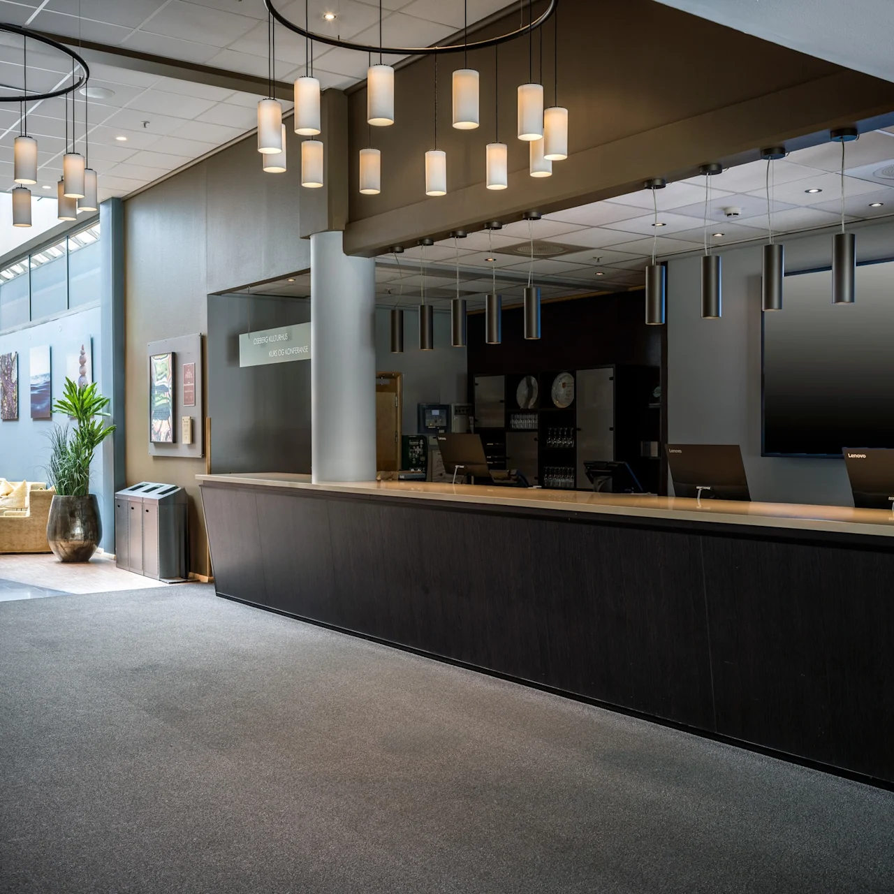 Lobbyreceptionsdisk, obemannad, med minimalistisk design. Taklampor hänger ovanför. Mysig sittgrupp med konstverk och krukväxter i närheten. Text: "Check out 12:00, Check in 15:00."