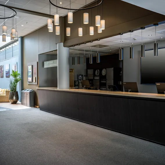 Resepsjonsdisk i lobby, ubemannet, med minimalistisk design. Taklamper henger over. Koselig sittegruppe med kunstverk og potteplanter i nærheten. Tekst: "Check out 12:00, Check in 15:00."