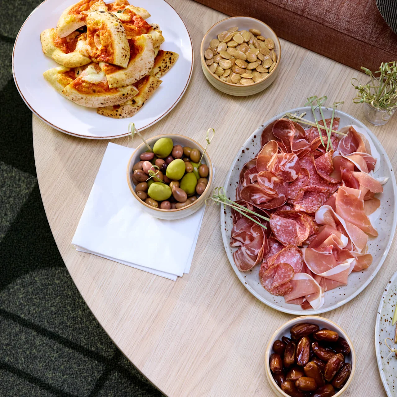 A variety of appetizers on a wooden table: a platter of cured meats, bowls of olives, almonds, dates, zucchini, cheese, and a plate of flatbreads with sauce.