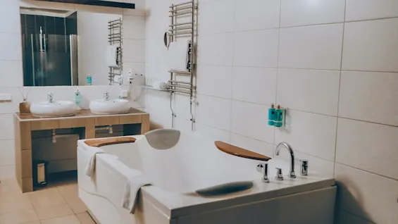 A white bathtub with wooden accents sits in a tiled bathroom. Two sinks and a large mirror are nearby, with a towel draped over the tub edge.