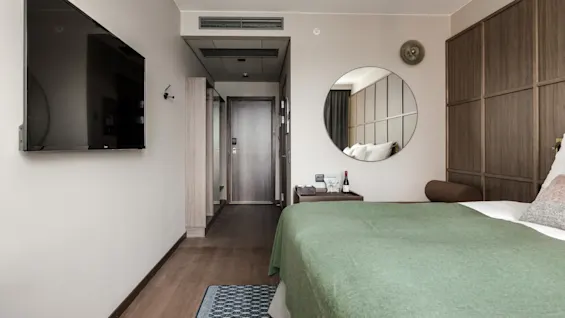 Et vægmonteret TV vender mod en seng med grønt sengetæppe ved siden af et rundt spejl, i et hotelværelse med træpanel og en lukket dør i baggrunden.
