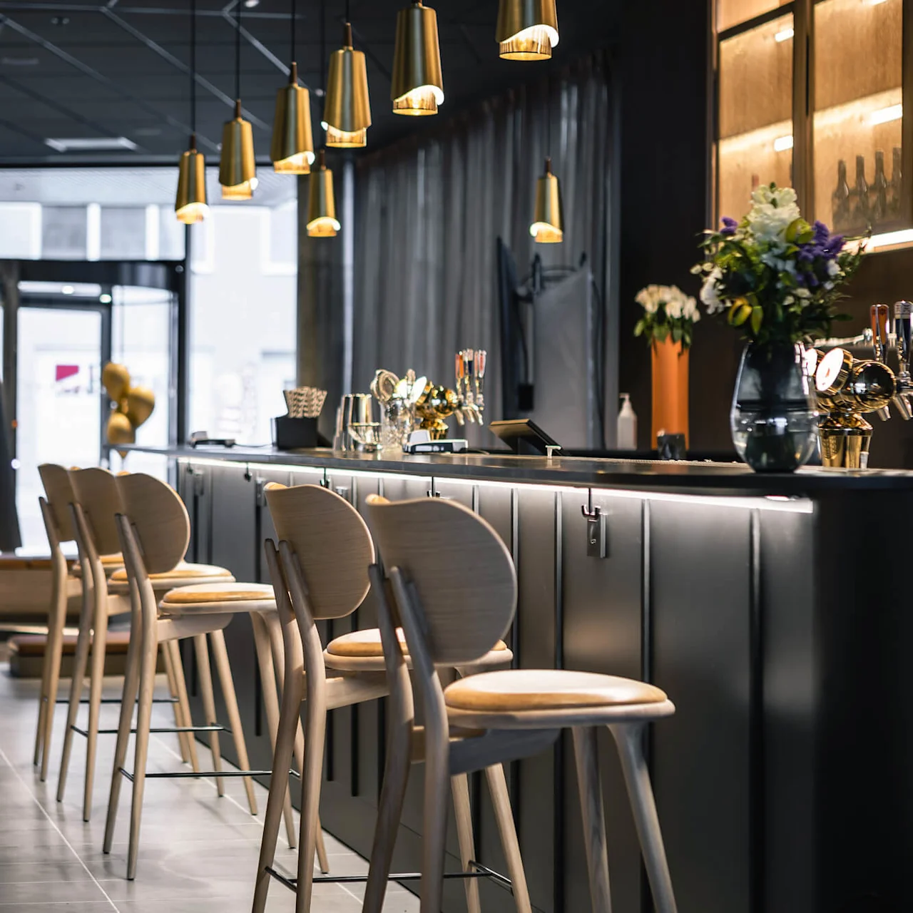 Four wooden bar stools stand at a sleek, illuminated bar in an elegant, modern restaurant. Overhead, golden pendant lights hang, adding warmth to the stylish interior.