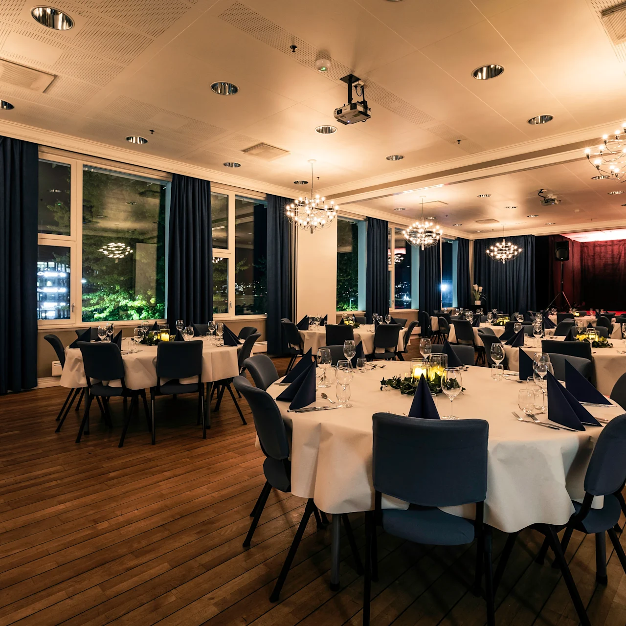 Elegant dining tables with white cloths and neatly folded napkins are arranged in a spacious, well-lit banquet hall, enhanced by chandeliers and surrounded by large windows and blue curtains.