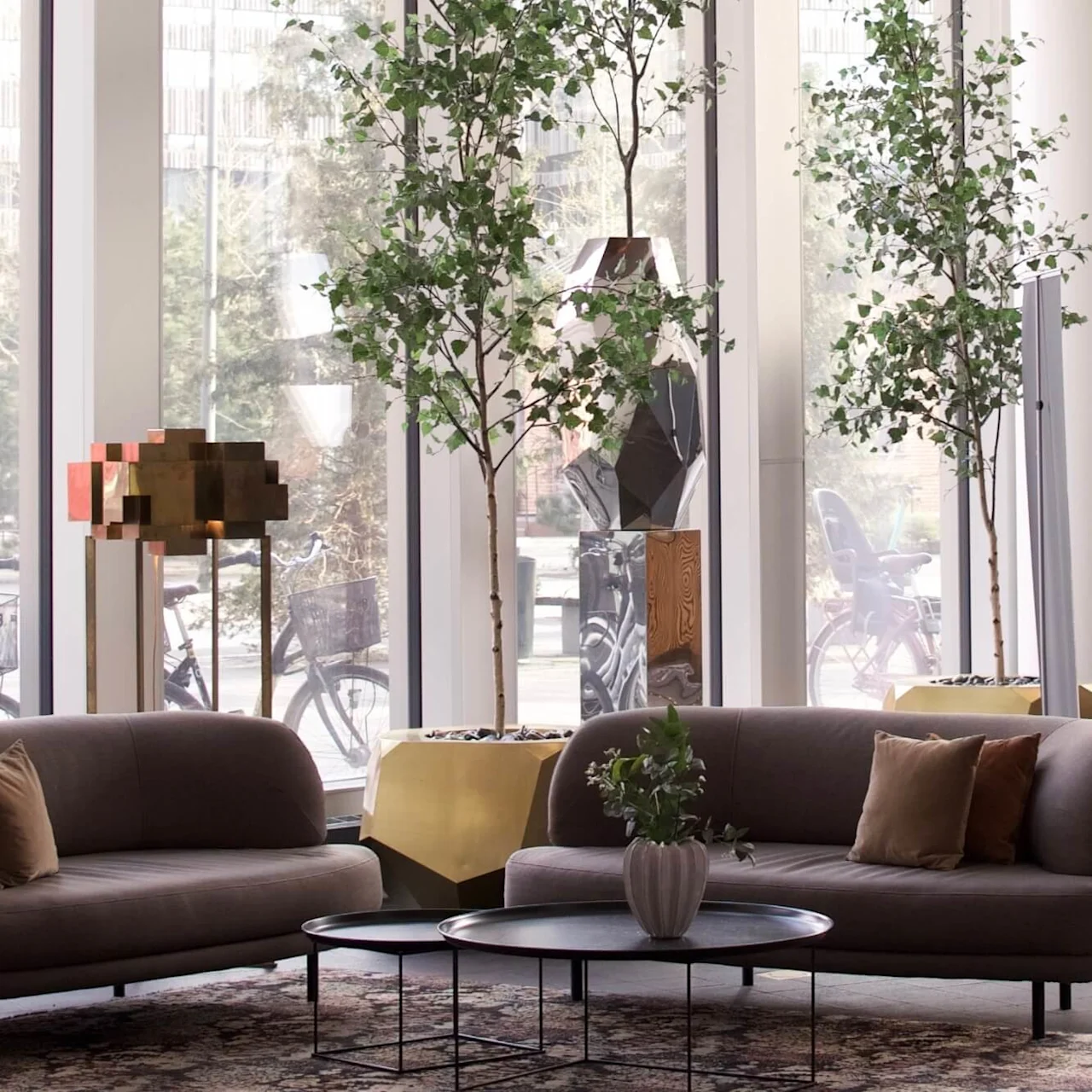 Sofaer med puter står mot hverandre, adskilt av runde bord, i en moderne lounge med høye planter og store vinduer som viser sykler utenfor.