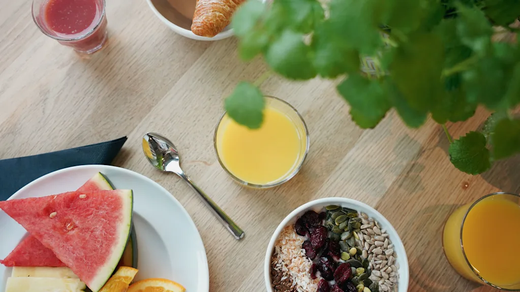 Aamiainen puupöydällä, jossa on croissantteja, vesimeloniviipaleita, kulho siemenillä ja marjoilla, lasilliset appelsiini- ja punamehua sekä vihreitä kasveja.