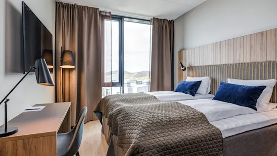 A bed with blue pillows and a brown quilt is placed by a window. Nearby, a desk with a chair and lamp is against the wall with a mounted TV.