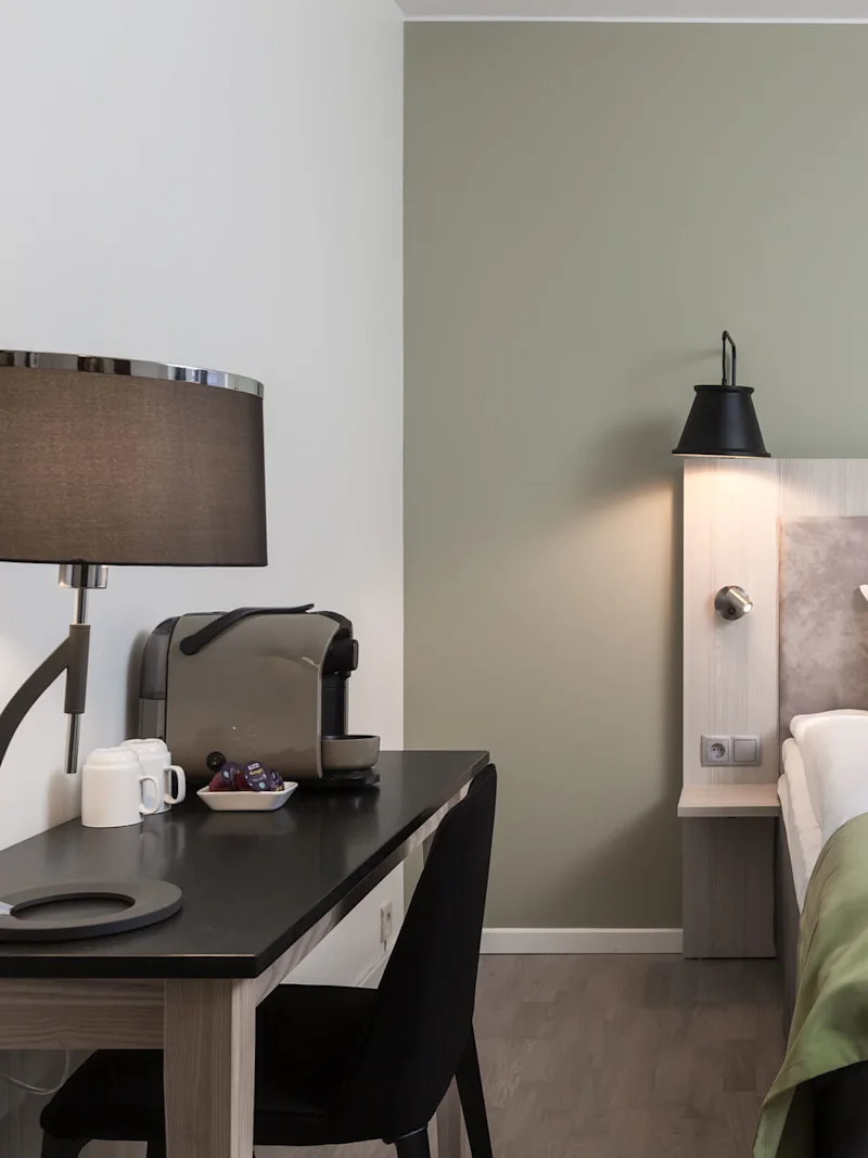 Siististi pedattu sänky, jossa on valkoiset tyynyt ja vihreä päiväpeite. Sivussa on pöytä, jossa on kahvinkeitin, kuppeja ja lamppu. Huone on moderni ja minimalistinen.