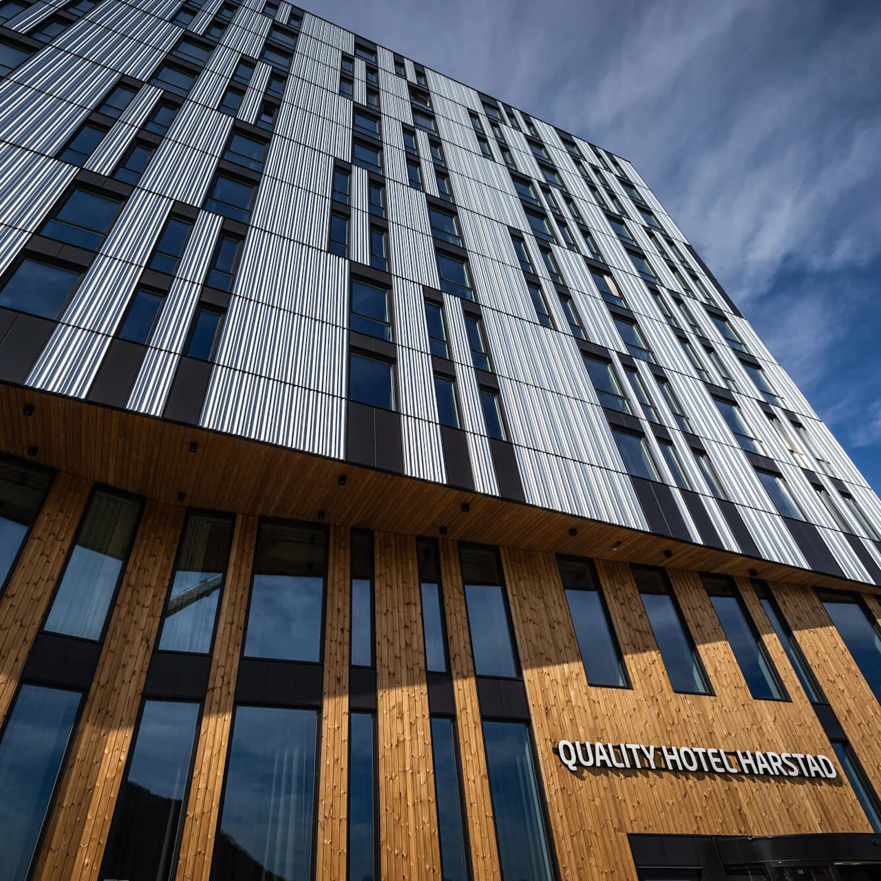En høj, moderne bygning med en geometrisk facade af metal- og træpaneler står under en blå himmel. Skiltet lyder "QUALITY HOTEL HARSTAD".