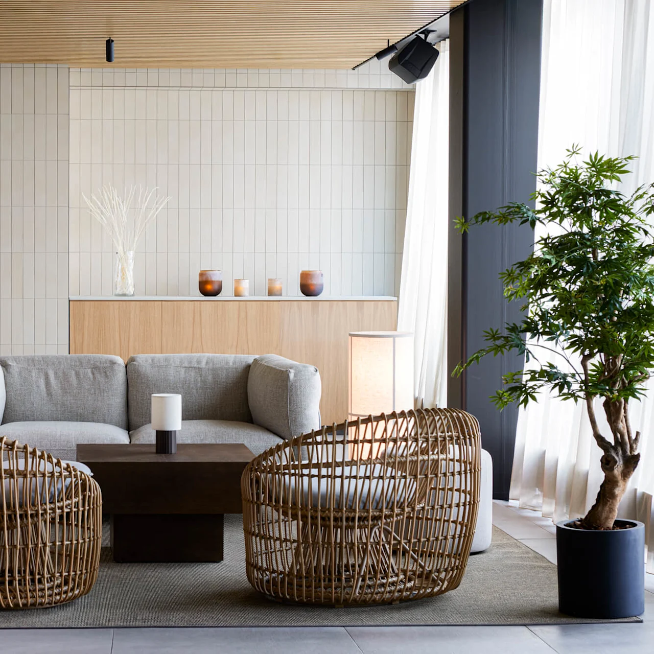 A gray sofa and two wicker chairs surround a dark coffee table in a modern living room. The setting includes potted plants, soft lighting, and light wooden accents.