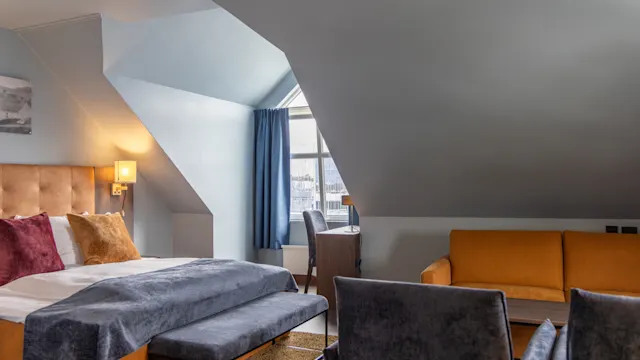 Et koselig soverom med en stor seng, polstret hodegjerde og fargerike puter ved siden av et vindu med blå gardiner. Ved siden av finnes en oransje sofa og et tre skrivebord.
