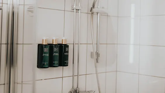Tre mørkegrønne flasker mærket 'shower gel', 'shampoo', 'conditioner' på en hylde, nær et brusehoved i et klinkebelagt badeværelse.