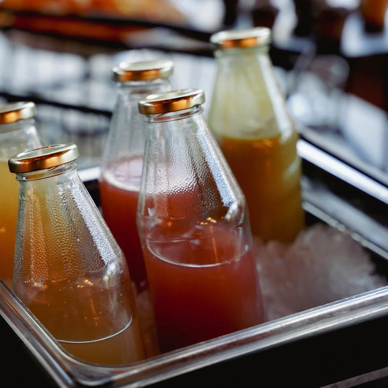 Glasflasker med metal kapsler, indeholdende orange og pink drikkevarer, køles på is i en metalbakke, antyder en bar- eller café-indstilling.