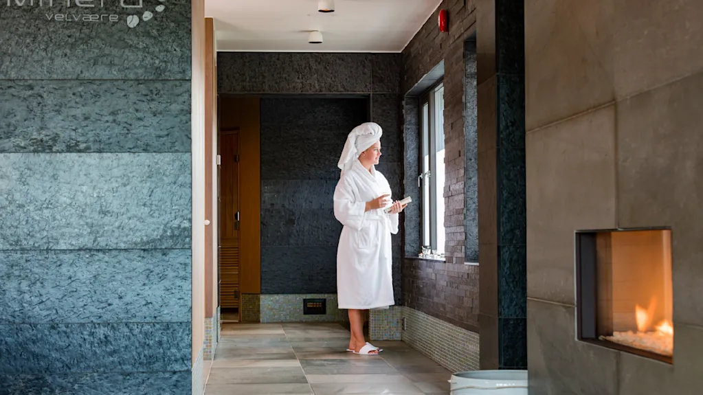 En person i badekåbe og håndklædehætte står ved et vindue i et spa, holder en kop, med en moderne pejs på en flisevæg i nærheden. Tekst: "minera velvære".