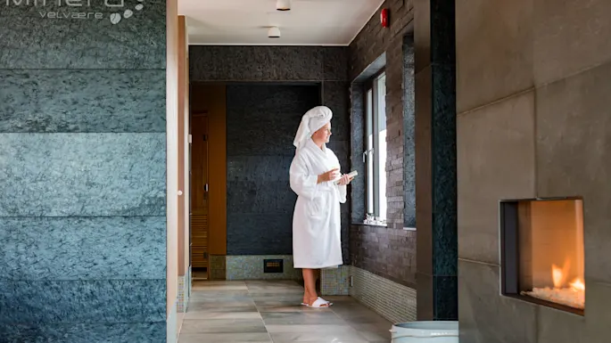 A person in a bathrobe and towel cap stands by a window in a spa, holding a cup, with a modern fireplace on a tiled wall nearby. Text: “minera velvære”.