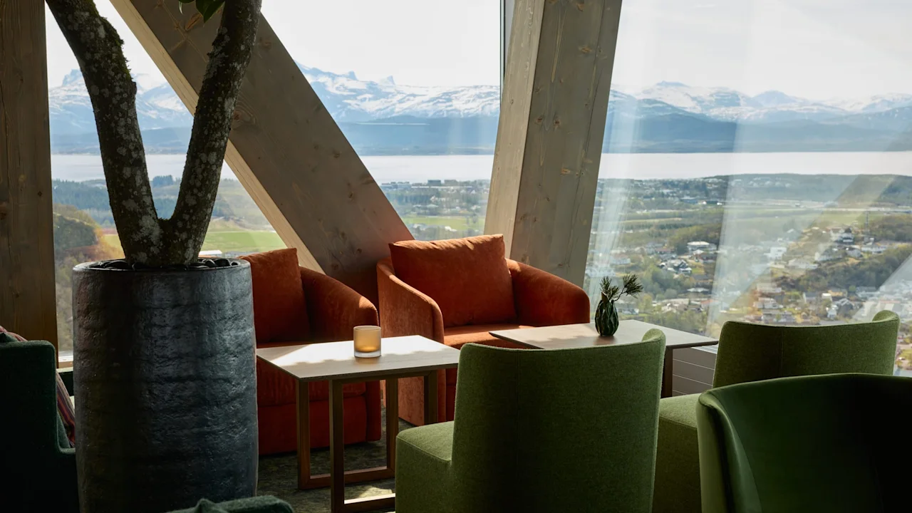 Cozy armchairs surround a small table by large windows, offering a scenic view of a fjord and mountains. A potted tree stands nearby, adding to the relaxing atmosphere.