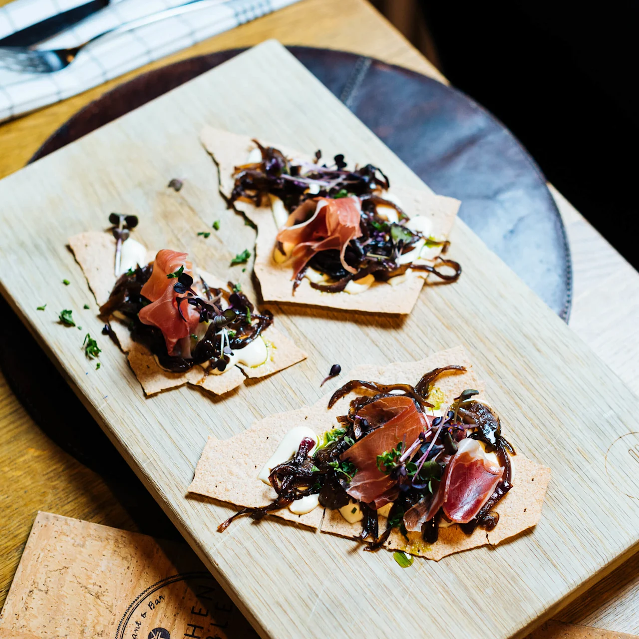 Frasiga flatbröd toppade med prosciutto, karamelliserad lök och mikrogrönt ligger på en träbricka, placerad på ett träbord med bestick och servetter i närheten.