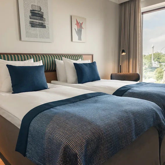 A bright hotel room with two comfortable beds, blue accents, and a view of the city from the window.