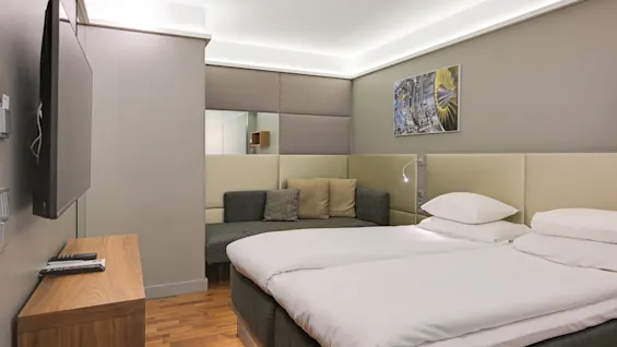 En modern hotellrum med en snyggt bäddad säng, intilliggande soffa och väggmonterad TV, i en minimalistisk design med neutrala färger och mjuk belysning.
