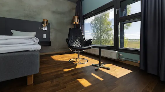 A modern hotel room with a stylish black chair facing a window, next to a bed with white sheets. Sunlight streams in, illuminating the wooden floor.