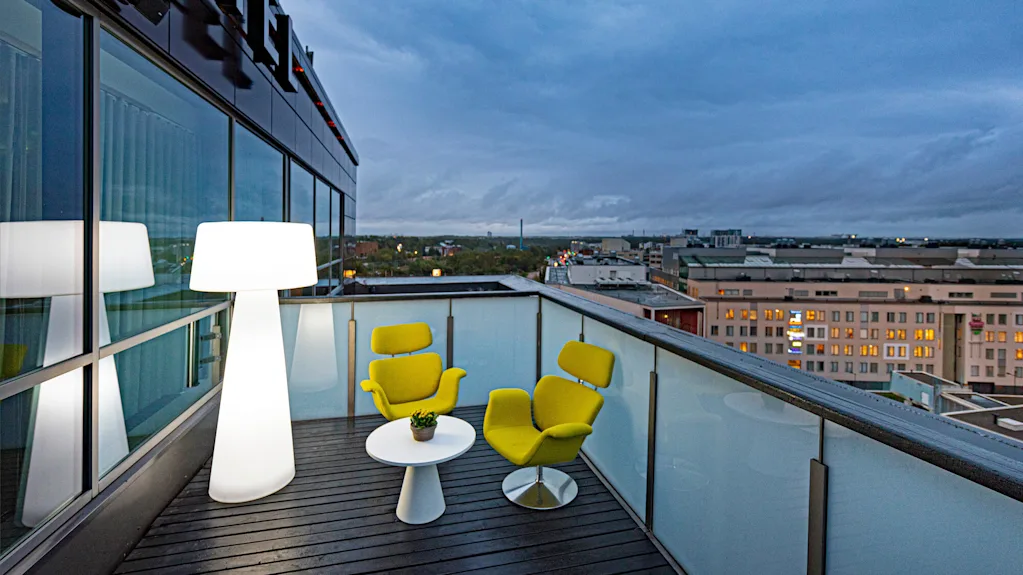 A rooftop terrace features two yellow chairs and a white table with a plant, illuminated by a tall modern lamp, surrounded by glass panels overlooking the city skyline.