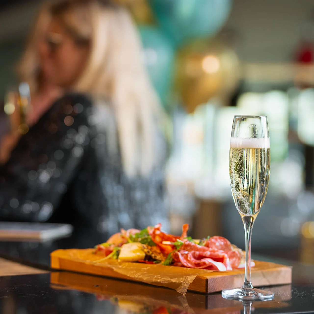 Ett glas champagne står på en svart disk bredvid en träplatta med olika sorters kött, med suddiga festdekorationer och människor i bakgrunden.