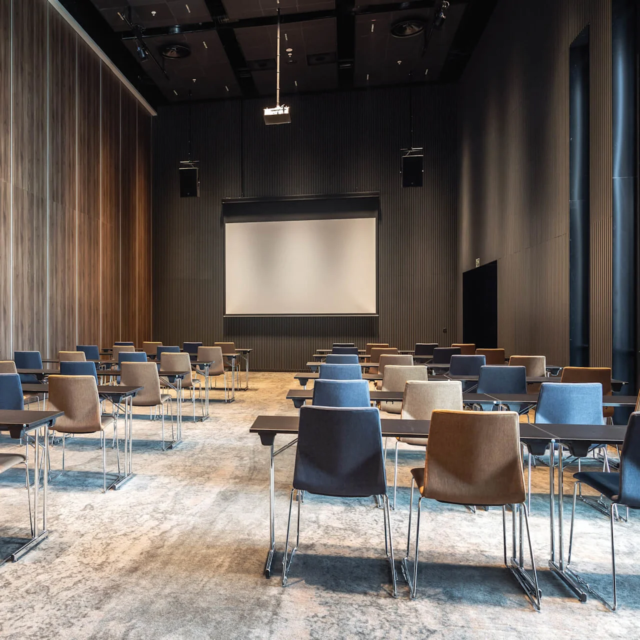 Rækker af tomme stole og skriveborde vender mod en stor projektorskærm i et rummeligt, moderne konferencelokale med vægge af træpanel og et tekstureret tæppe.